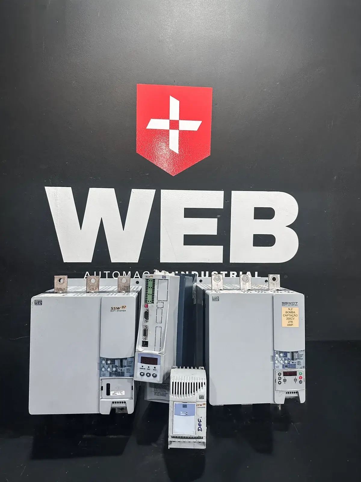 Inversores de frequência e soft starters WEG na bancada, diante do logo da WEB Automação Industrial
